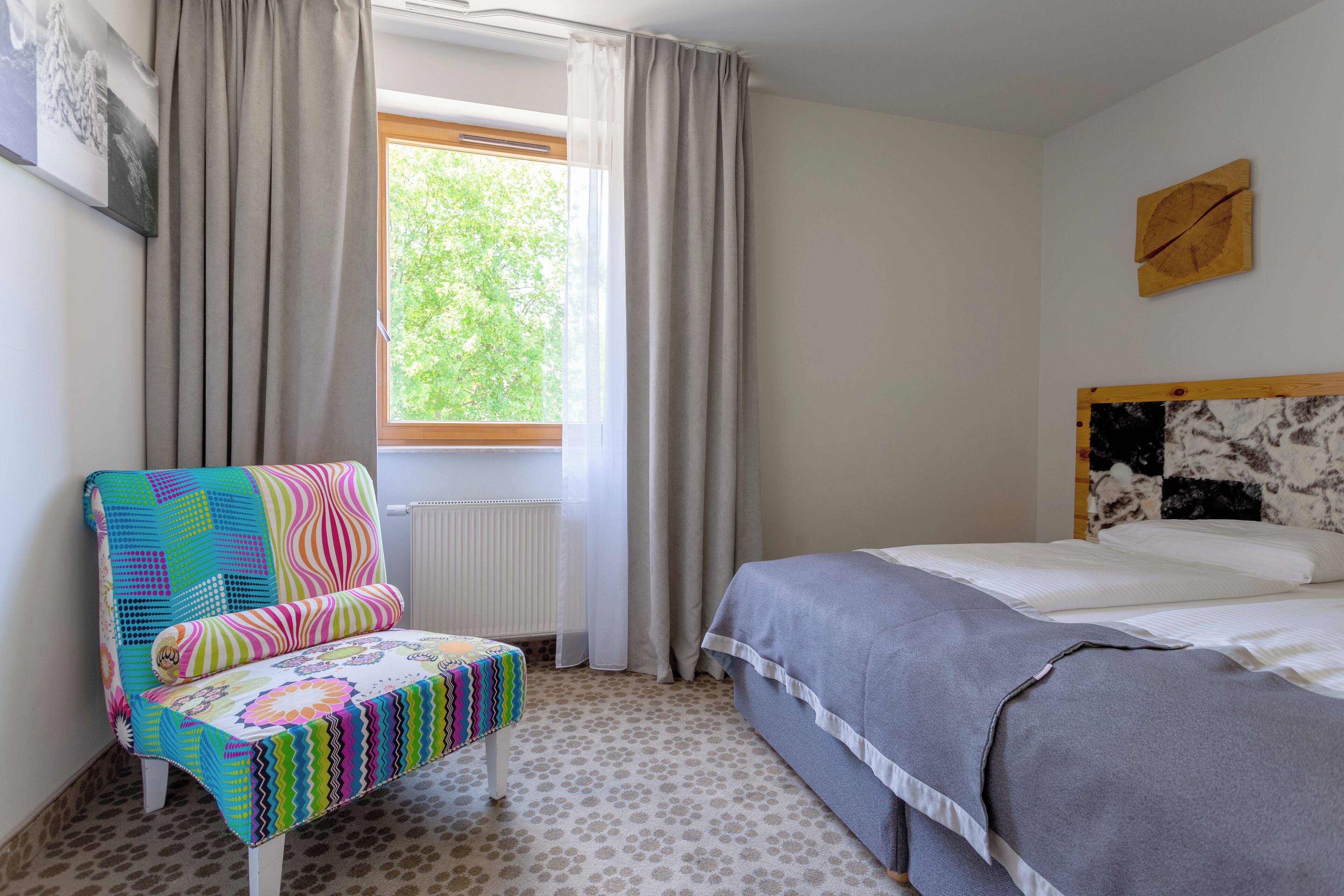 A bedroom with a bed, a colorful chair, a window with curtains, and a radiator.