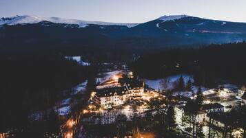 A snow-covered mountain landscape with a lit-up building complex in the foreground, surrounded by trees and a small lake.