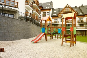 Un parque infantil con tobogán y estructuras para escalar, rodeado de una zona de grava y edificios residenciales al fondo.
