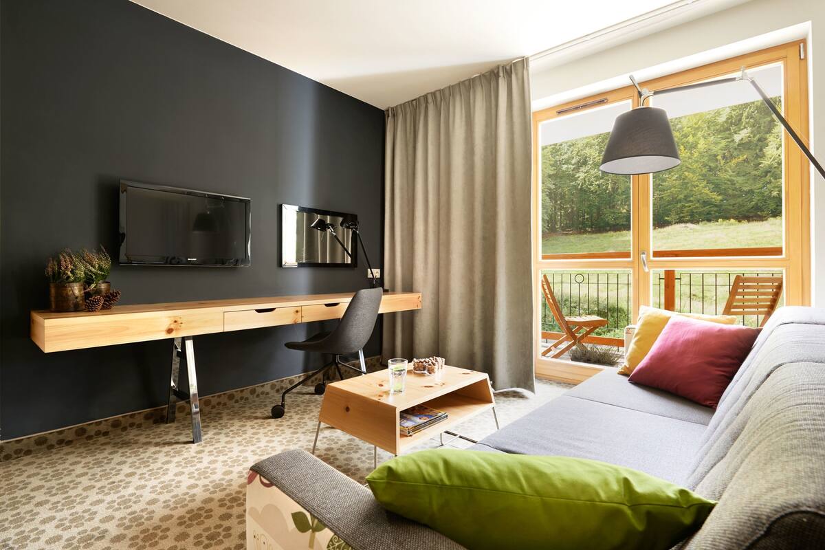 Une chambre moderne équipée d’un bureau, d’une chaise et d’un canapé, d’une télévision et d’une fenêtre donnant sur un paysage verdoyant.