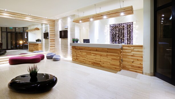 A modern reception area with a wooden reception desk, seating area with a pink sofa and blue chairs, and a black decorative planter.