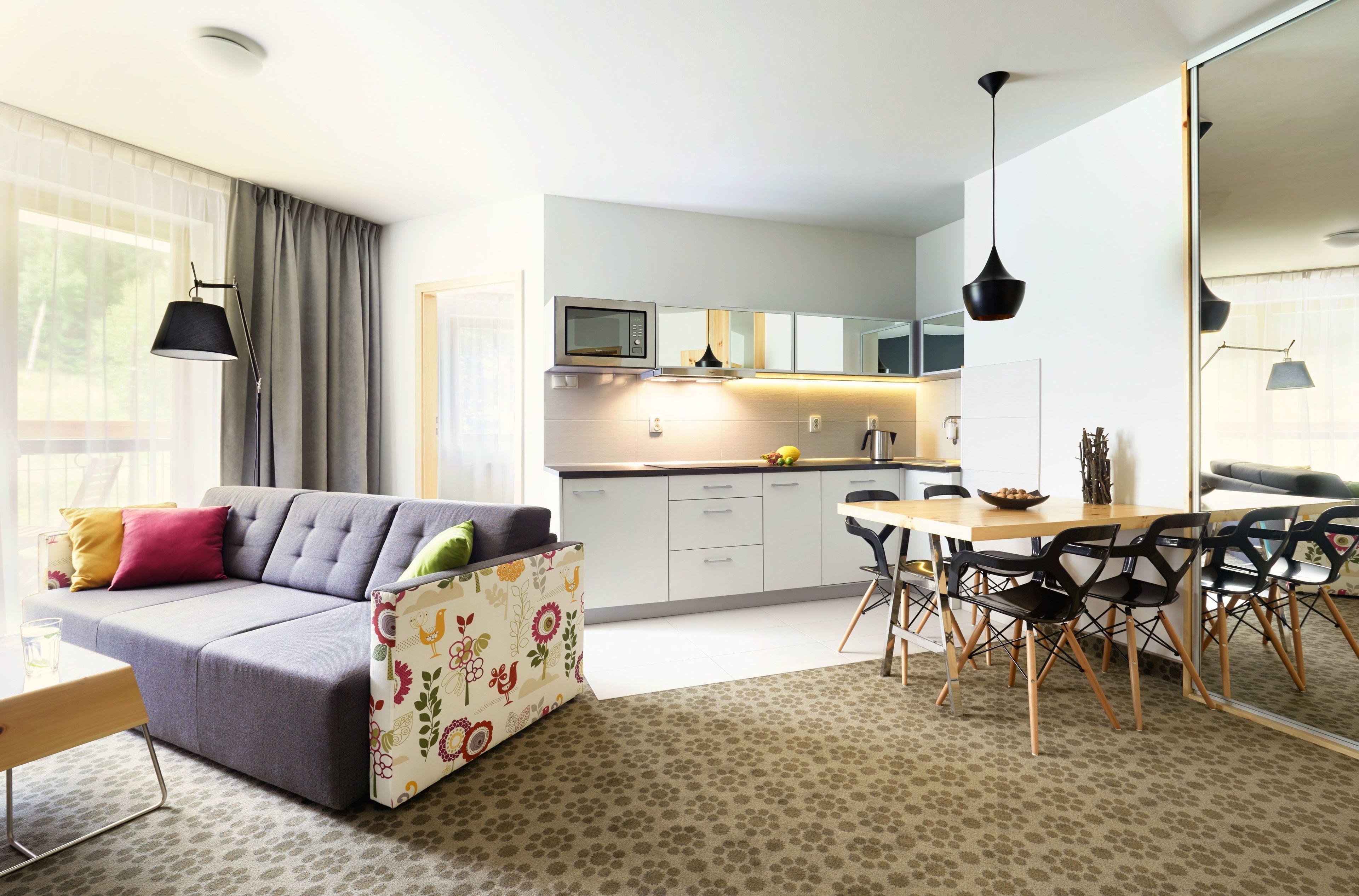A modern living room with a sofa, a dining table with chairs, a kitchen area with cabinets and a microwave, and a large mirror.