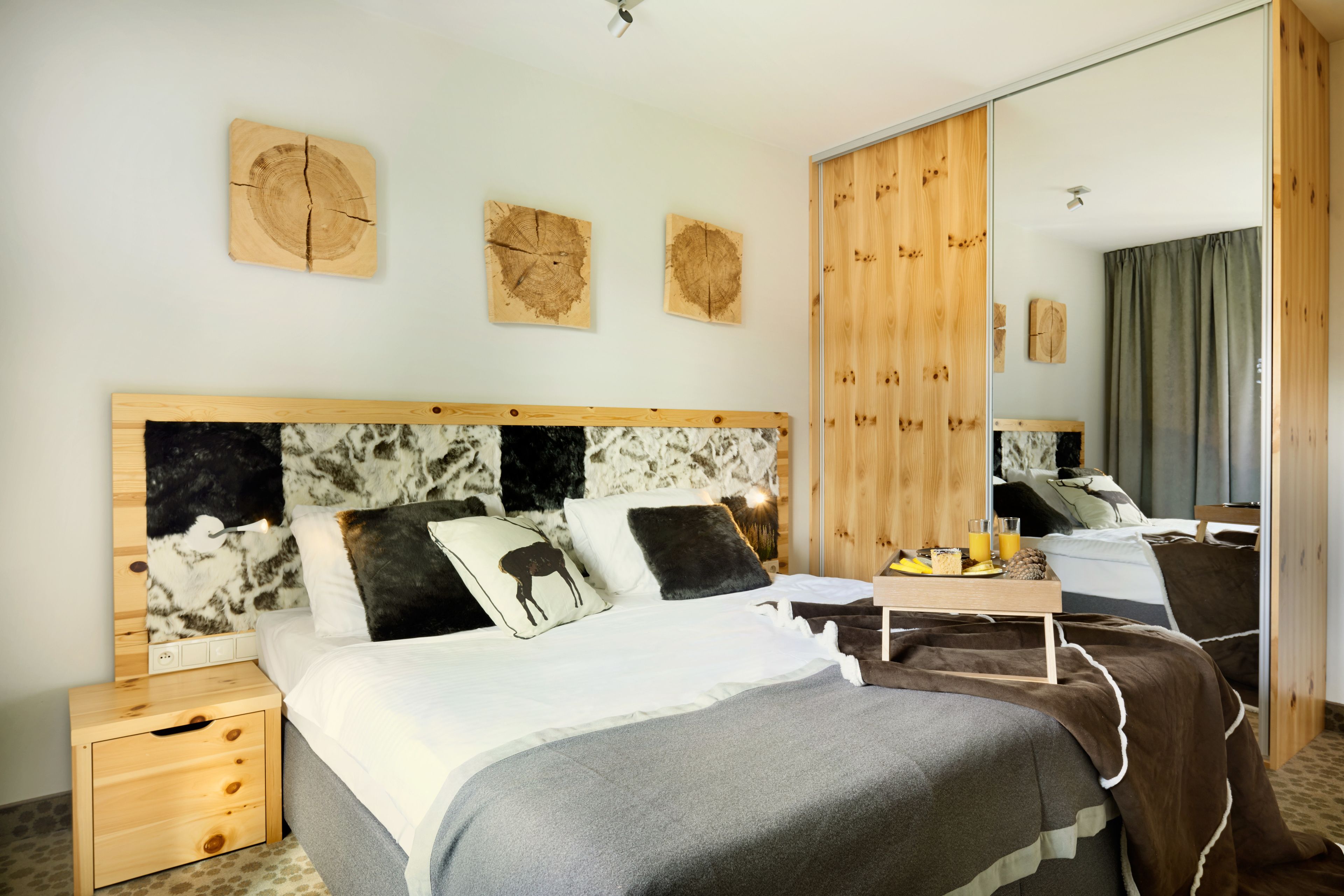 A modern hotel room with a wooden headboard, a large mirror, and a bed with a grey and brown blanket.
