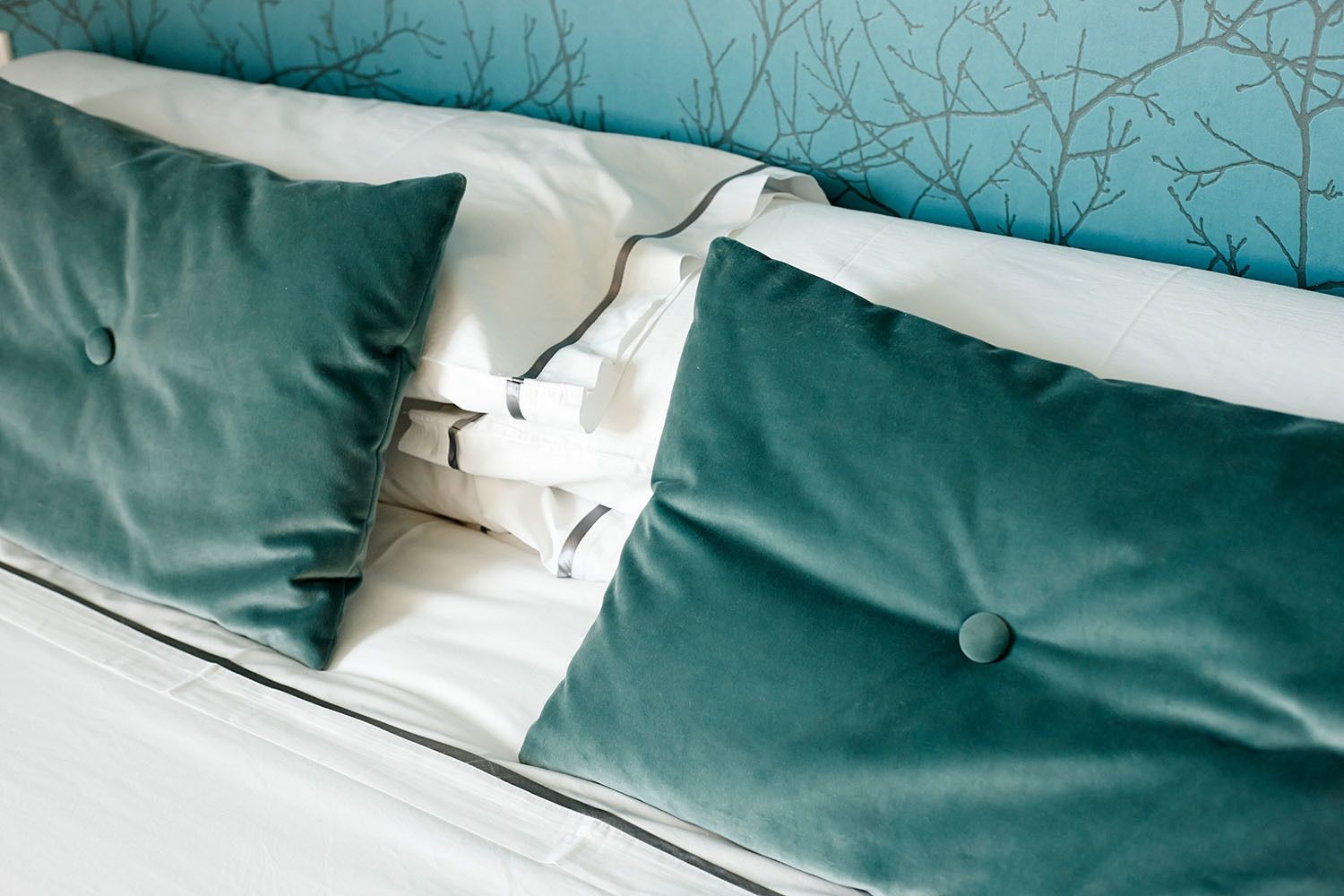 Cama con almohadones color turquesa y ropa de cama blanca, frente a una pared azul con diseño de ramas de árbol.