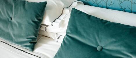 Cama con almohadones color turquesa y ropa de cama blanca, frente a una pared azul con diseño de ramas de árbol.