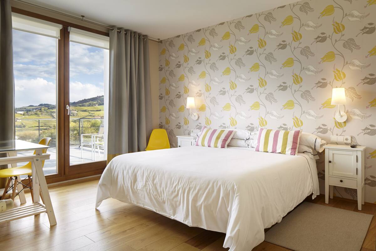 Een slaapkamer met een groot bed, een bureau en een stoel. Er is een raam met gordijnen, een nachtkastje met een lamp en een wand met een bloemenmotief.