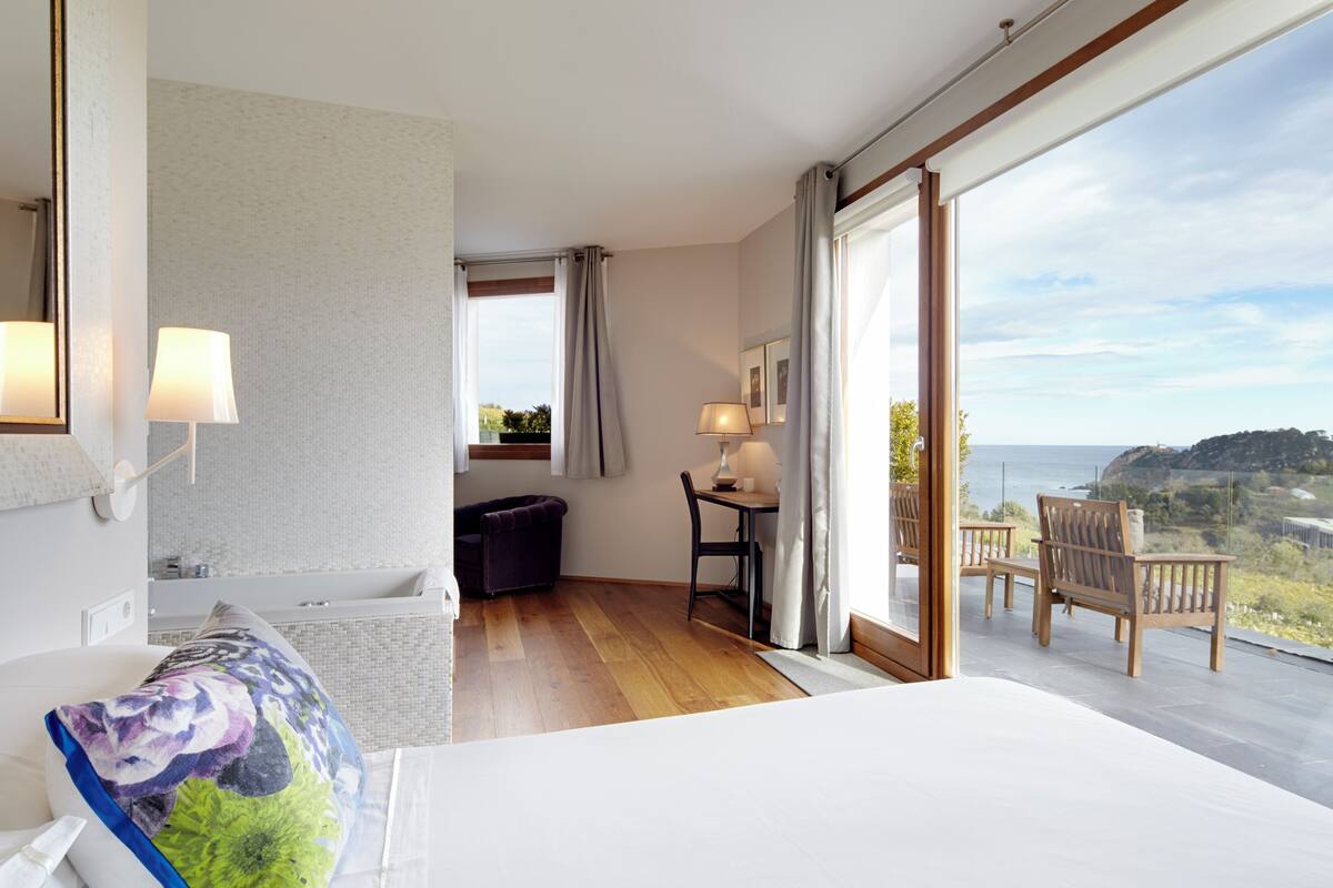 Een hotelkamer met een groot bed, een bureau met stoel, een bijzettafeltje met lamp, uitzicht op het omliggende landschap en een balkon met stoelen.