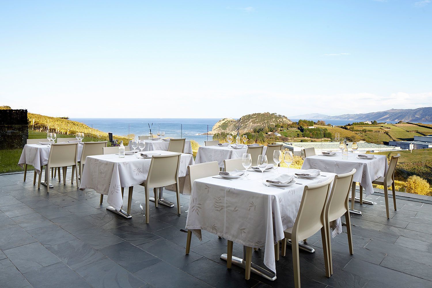 Un restaurante con terraza al aire libre, con vistas panorámicas al mar y colinas. Las mesas están cubiertas con manteles blancos y sillas.