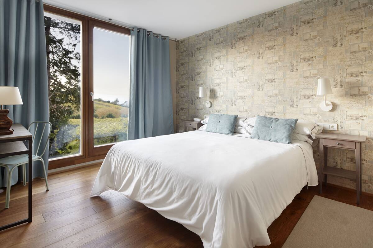 Een slaapkamer met een groot bed, een bureau en een stoel. Er is een raam met gordijnen, een wand met behang en twee nachtkastjes met lampen.