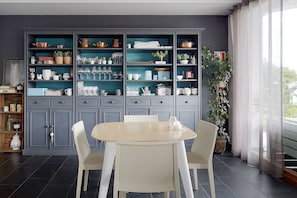 Comedor con mesa redonda y sillas, un mueble empotrado con utensilios de cocina y un ventanal con cortinas.