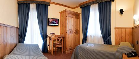 A hotel room with two single beds, a wooden wardrobe, a desk with a chair, and a television on the wall.