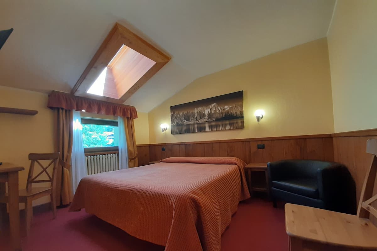 A bedroom with a large bed, a wooden headboard, a wooden desk, a chair, a small table, a window with curtains, and a painting on the wall.