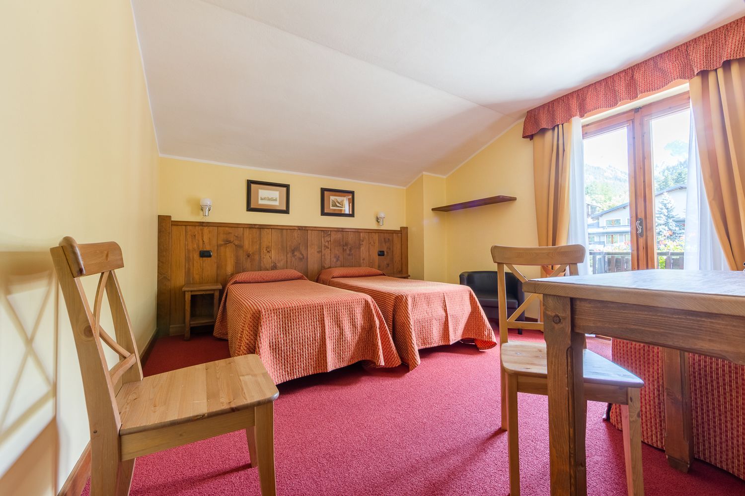 A room with two beds, a wooden table, and a chair. There are two framed pictures on the wall.