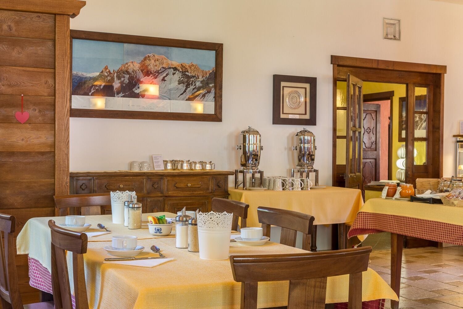 Un comedor con mesas y sillas de madera, un espacio tipo bufé con cubiertos, y un cuadro de un paisaje montañoso en la pared.