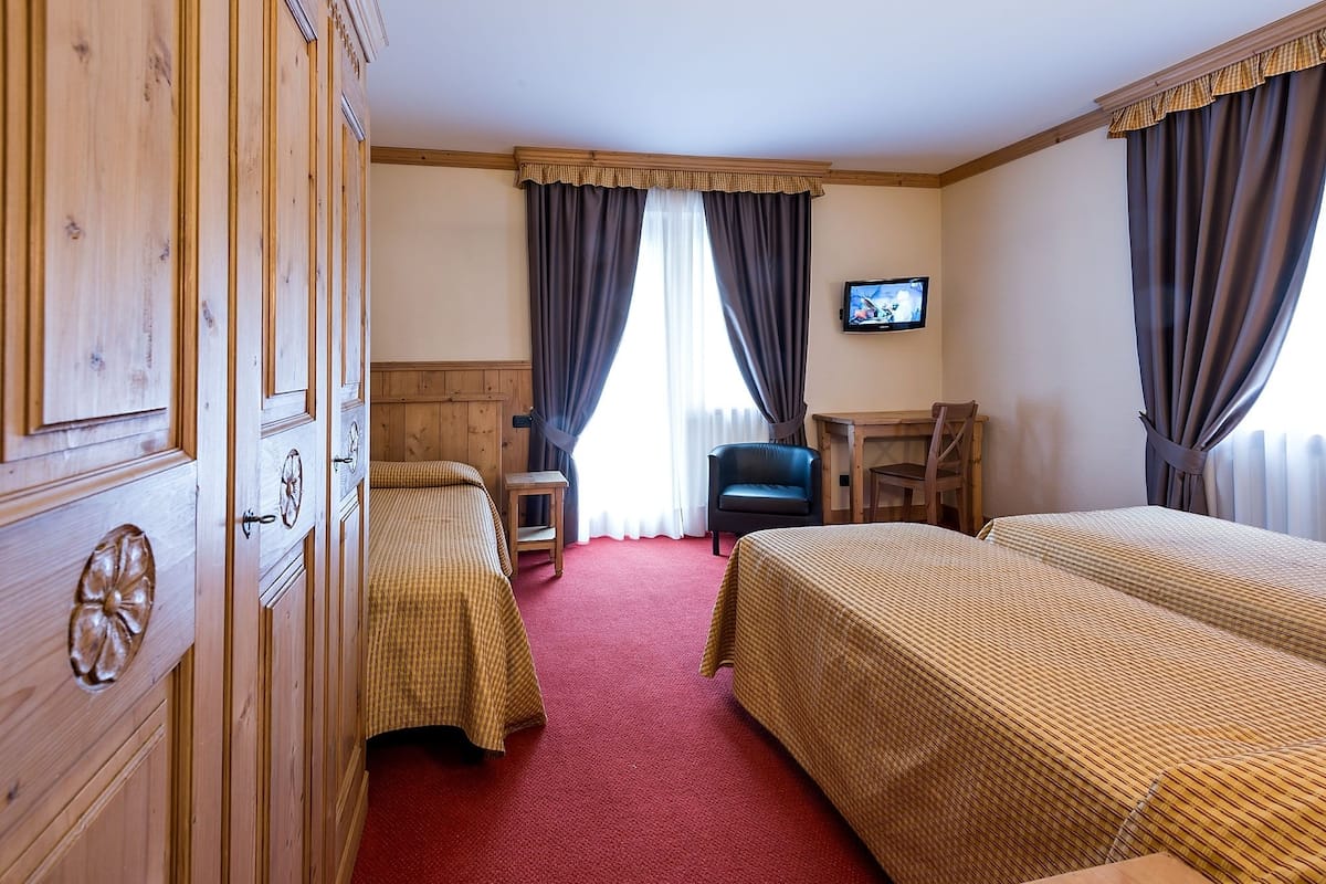 A hotel room with two beds, a wooden wardrobe, a small table, a chair, and a television on the wall.