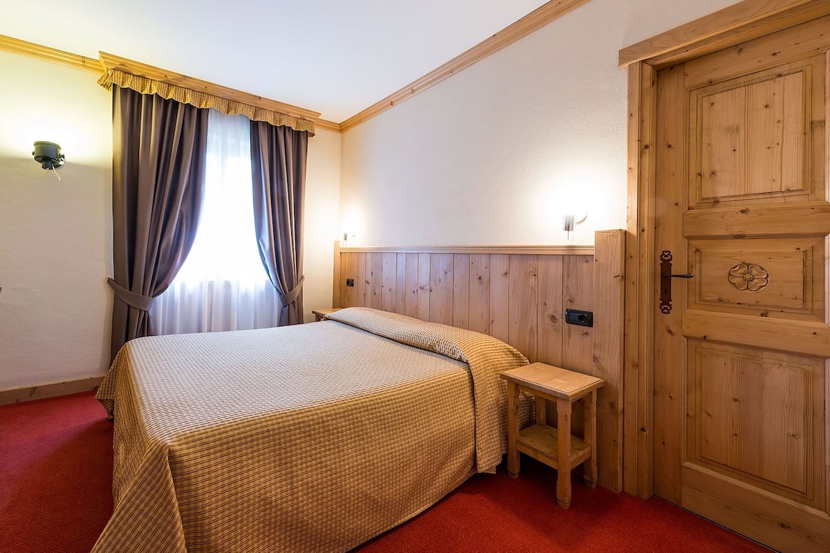 A hotel room with a bed, a wooden nightstand, a small table, a wooden door, and a window with curtains.