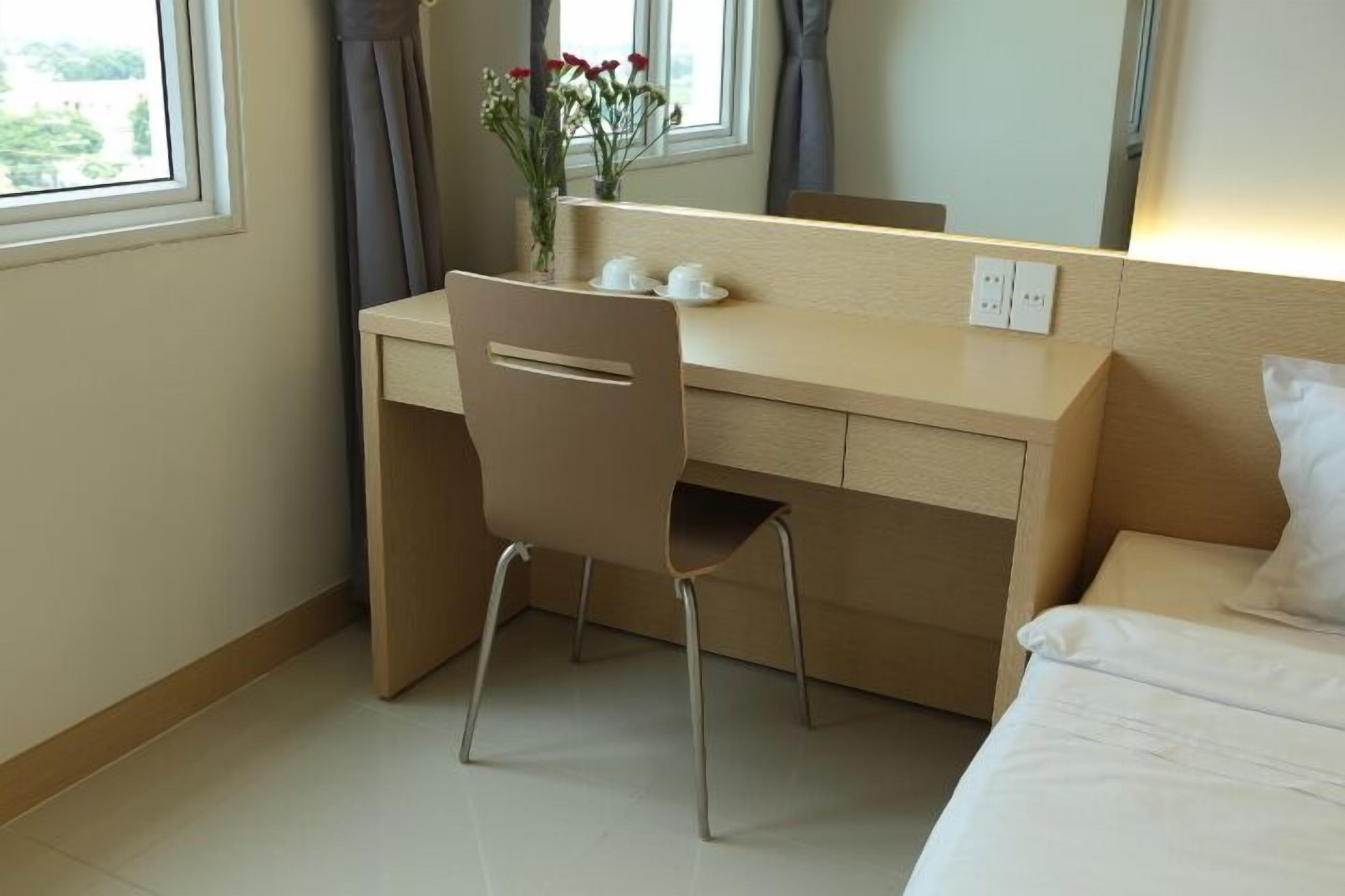 Ett hotellrum med ett skrivbord i trä, en stol, en säng med vita sängkläder och ett fönster med gardiner.