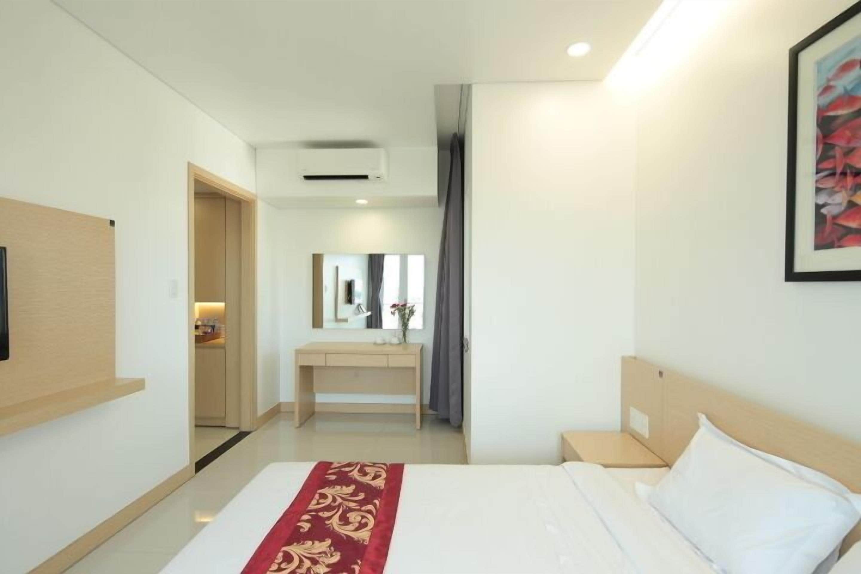 A modern hotel room with a large bed, a bedside table, a desk with a chair, a mirror, and a framed picture on the wall.