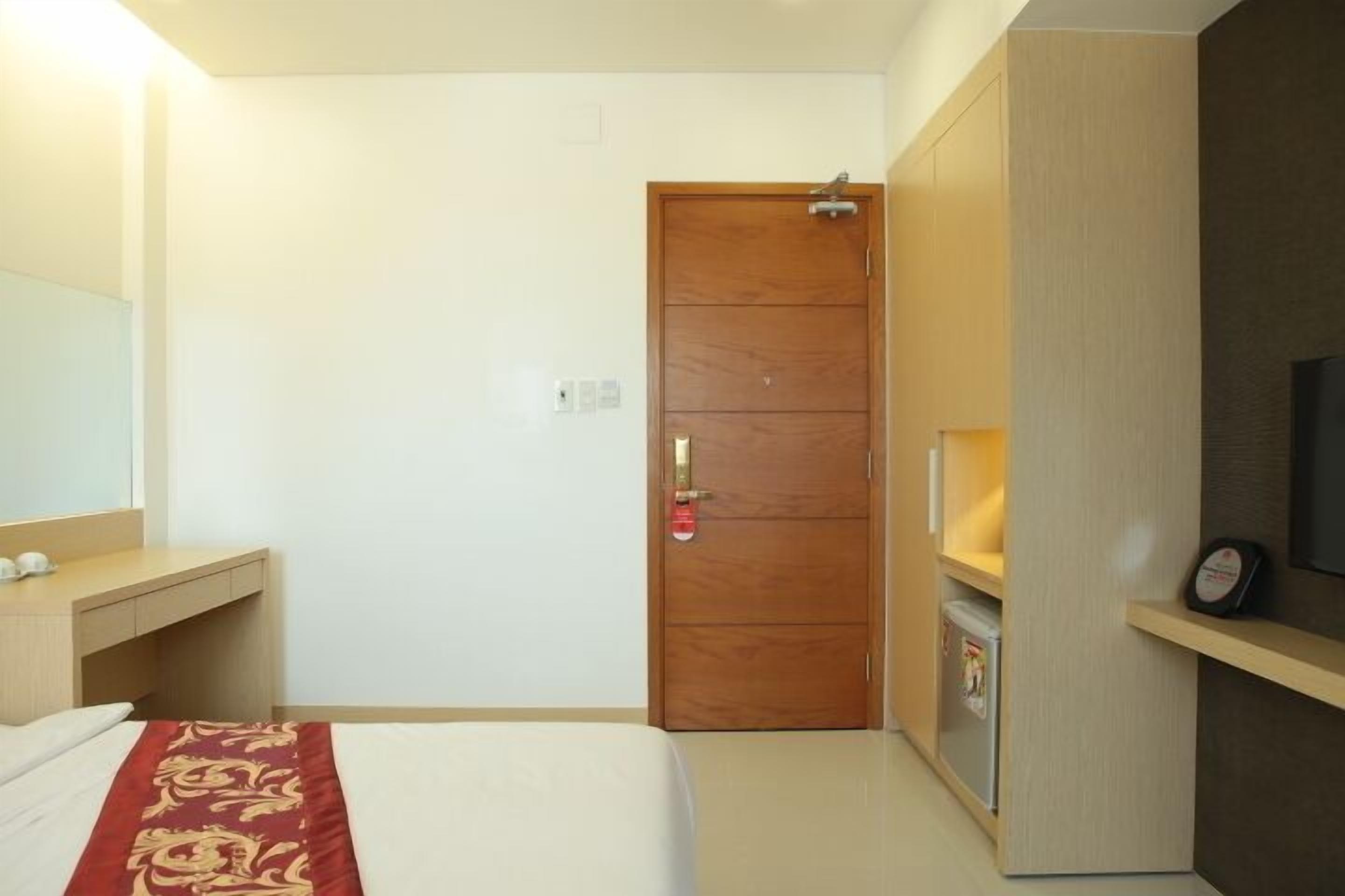 Habitación de hotel con una cama, un armario de madera, un televisor sobre un mueble bajo y una pequeña estantería con un reloj.