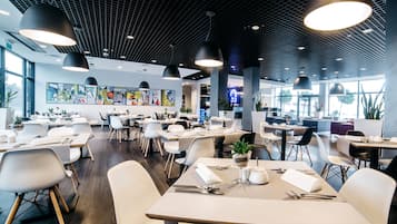 A modern restaurant with a grid-patterned ceiling, contemporary furniture, and large windows allowing natural light.