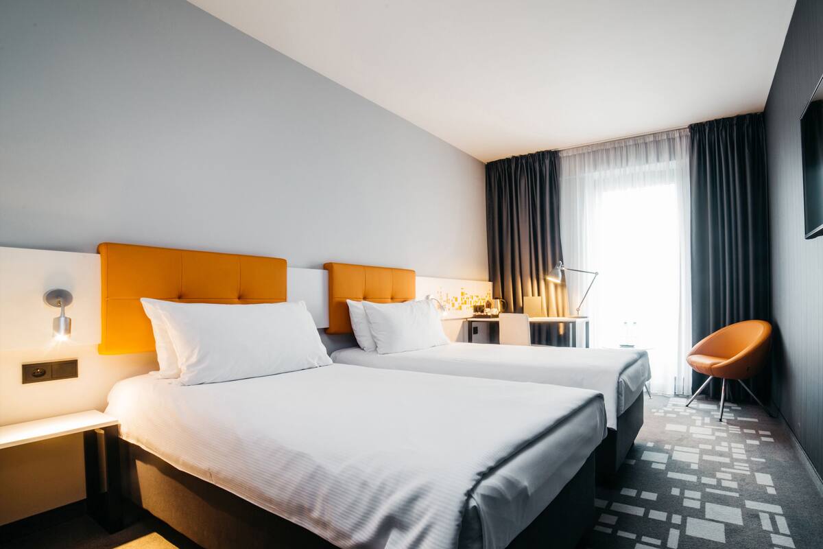 A hotel room with two beds, a desk, a chair, and a large window with curtains.