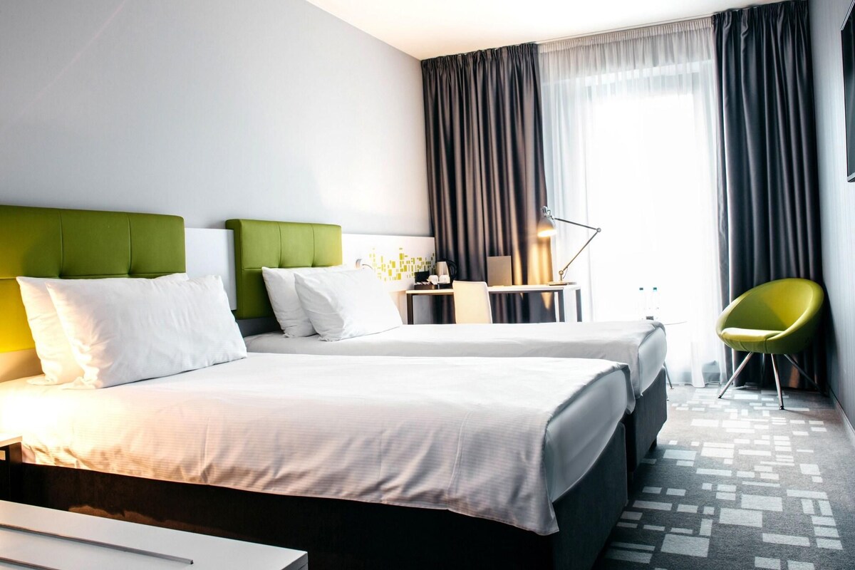 A hotel room with two beds, a desk, a chair, and a lamp.