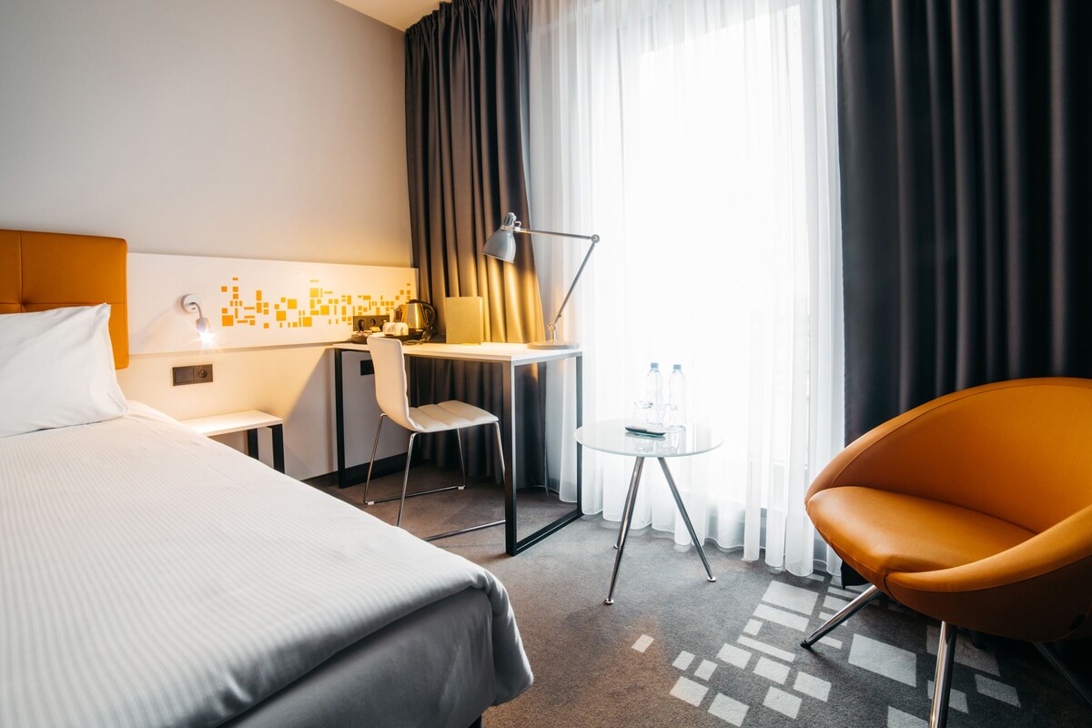 A hotel room with a bed, a desk, a chair, a lamp, and a small table with water bottles.