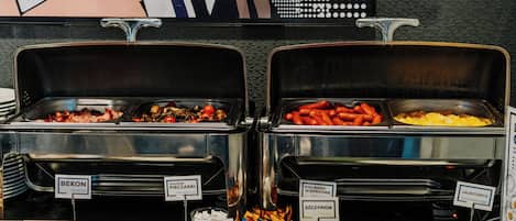 Un bufé con secciones de comida identificadas, que incluye tocino, champiñones fritos, kielbaski, huevos revueltos y cebollín.