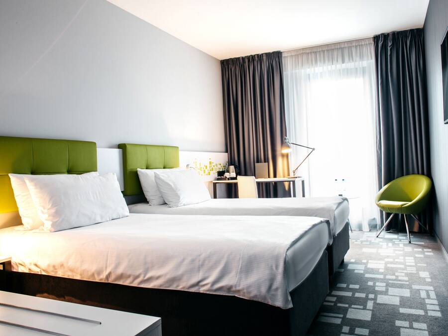 A hotel room with two beds, a desk, a chair, and a large window with curtains.