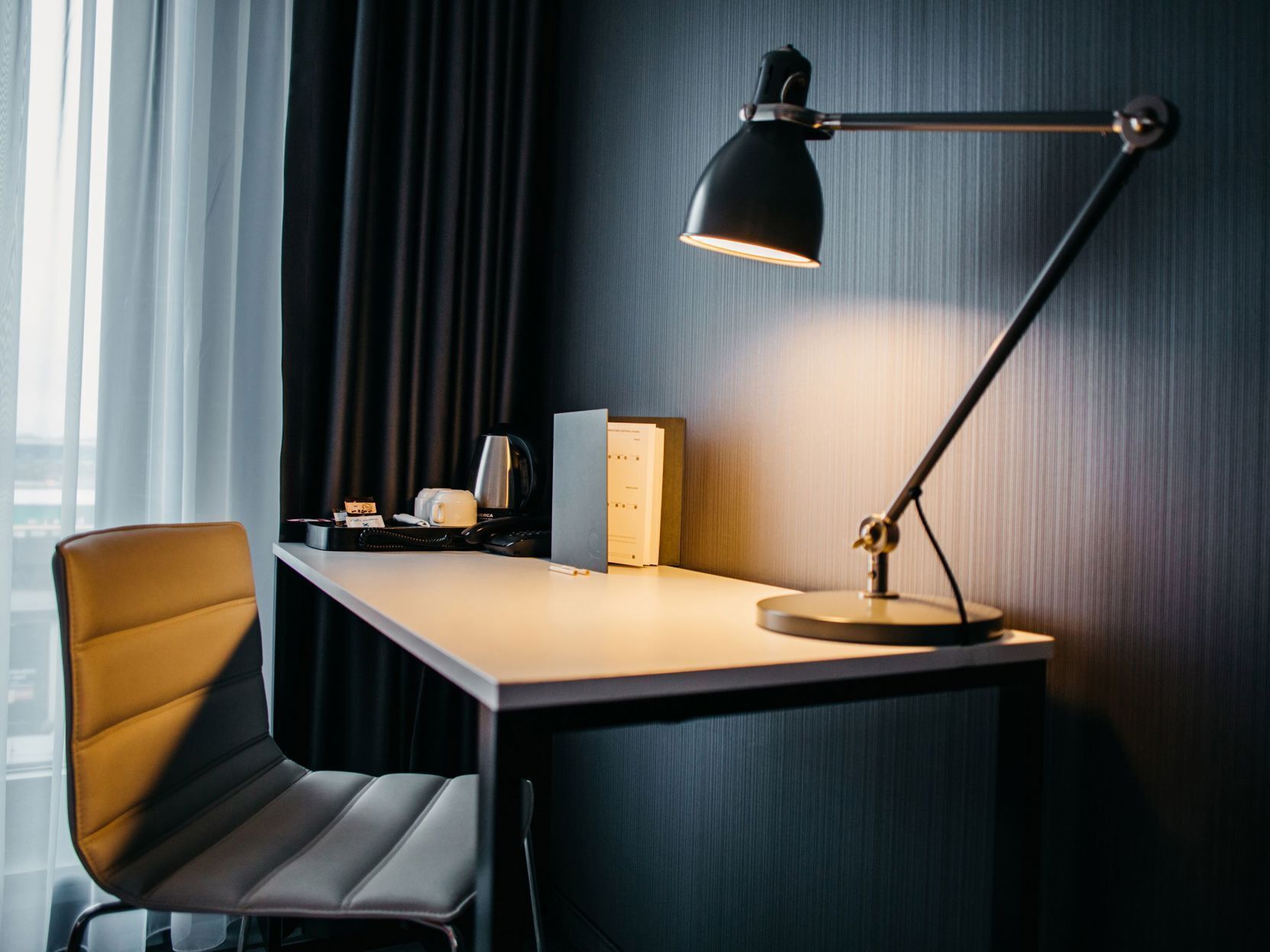 A modern hotel room with a desk, a chair, a desk lamp, a coffee maker, and a telephone.