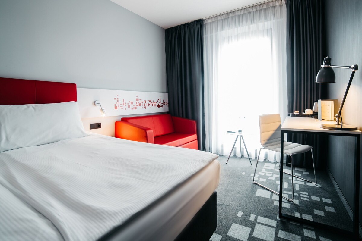 Une chambre d’hôtel avec un lit, un canapé rouge, un bureau avec une chaise, une lampe et une vue sur l’extérieur.