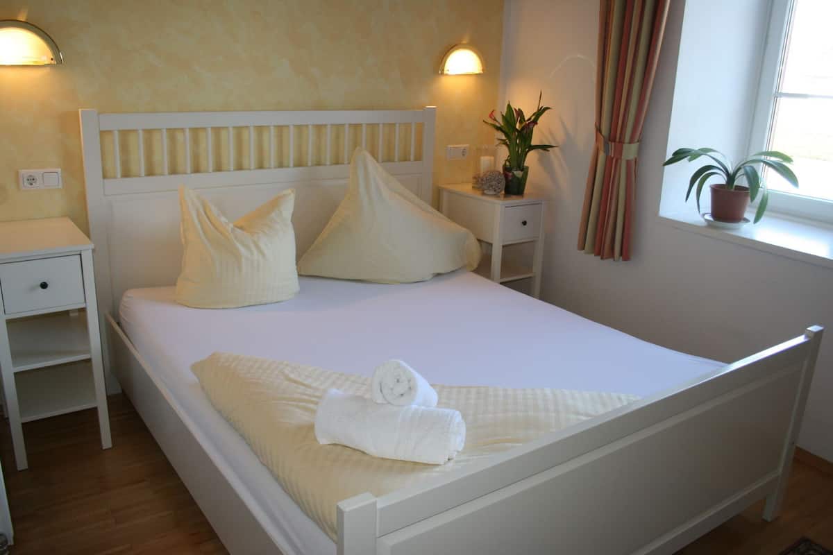 A neatly made bed with pillows, a bedside table, a nightstand, a potted plant, and a window with curtains.