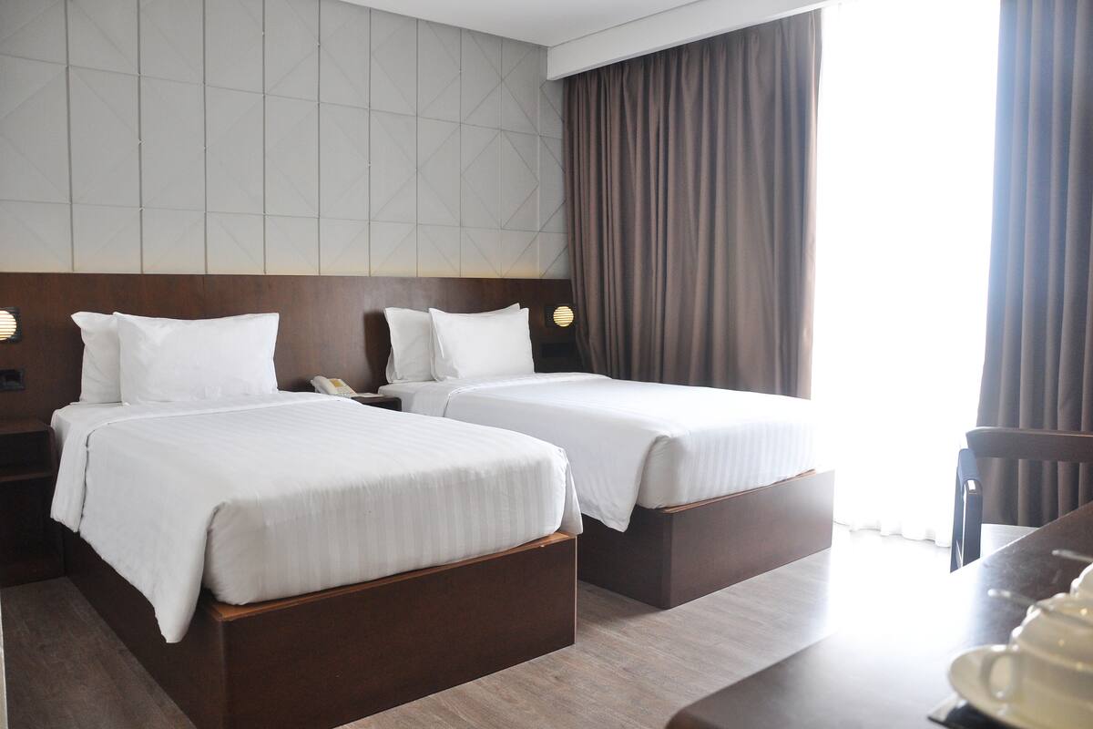 A hotel room with two beds, a wooden headboard, a bedside table with a lamp, and a view of the outdoors.