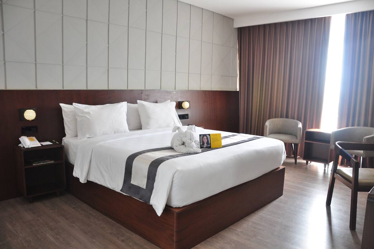 A hotel room with a large bed, a bedside table with a phone and a lamp, a chair, and a view of the outside through a window with curtains.