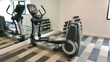 A gym with an elliptical machine, a set of dumbbells, and a television on the wall.