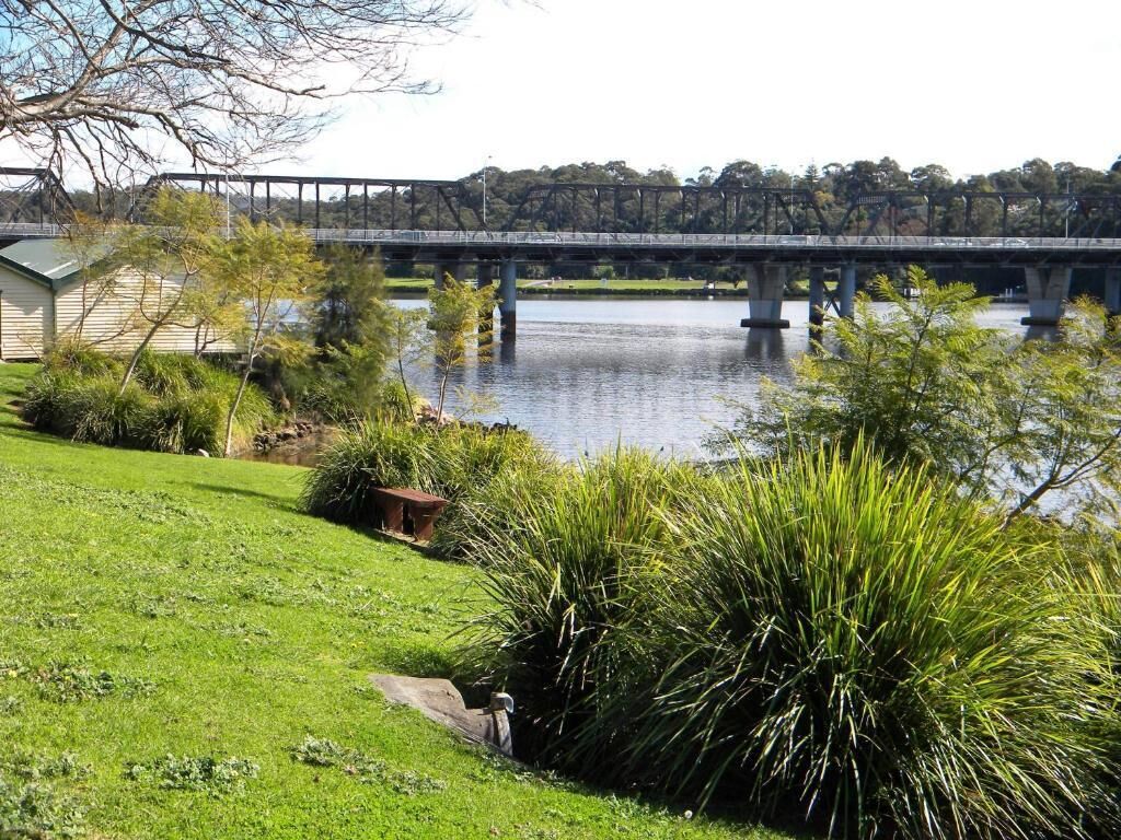 A bridge over a calm body of water, surrounded by lush greenery and a grassy area.