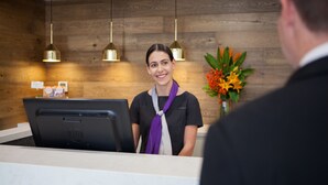 A hotel reception desk with a wooden wall background, a black HP computer, and a vase with orange flowers. - Quest Nowra (Nowra)