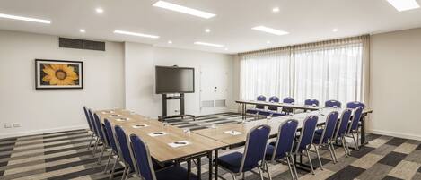 A conference room with a long table, blue chairs, a flat-screen TV, and a large window with curtains.