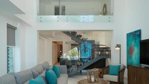 A spacious living area with a grey sofa, teal cushions, a wooden coffee table, and a TV on a wooden cabinet. A glass staircase leads to an upper level with a glass railing. - Gansevoort Dominican Republic (Sosua)