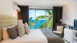 A hotel room with a large bed, a TV, a white dresser, and a balcony with a view of the ocean and palm trees. - Gansevoort Dominican Republic (Sosua)