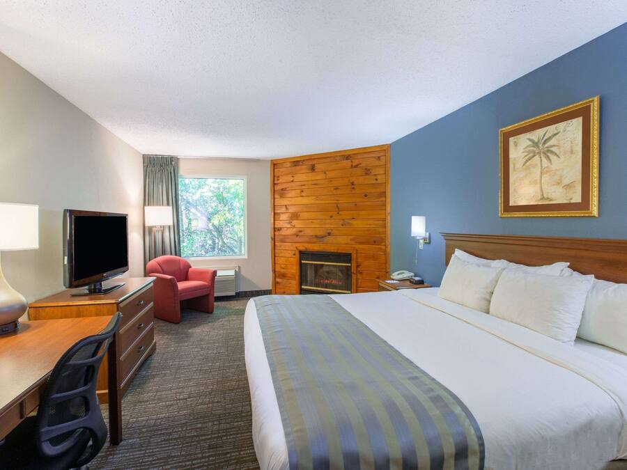 A hotel room with a bed, a desk with a computer, a television, a red chair, a fireplace, and a window with a view of trees.