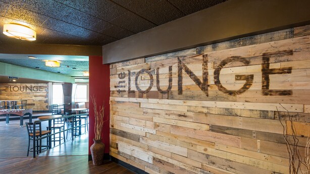 Pared de madera con la palabra "LOUNGE" en letras grandes, una zona de estar con mesas y sillas, y un letrero visible que indica el acceso al lounge.