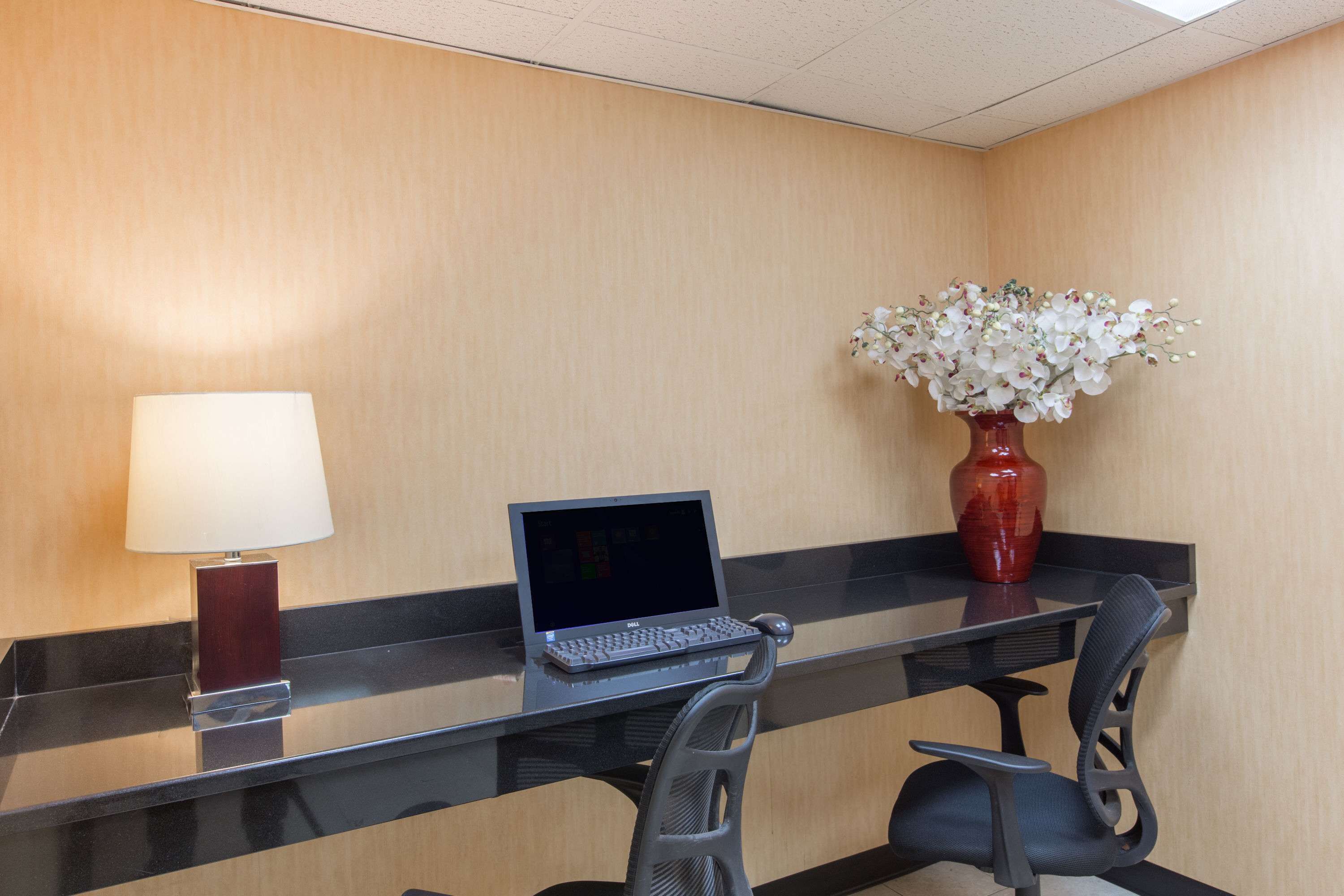 Une chambre d’hôtel avec un bureau en verre, un ordinateur portable, un vase rouge contenant des fleurs blanches et une lampe.