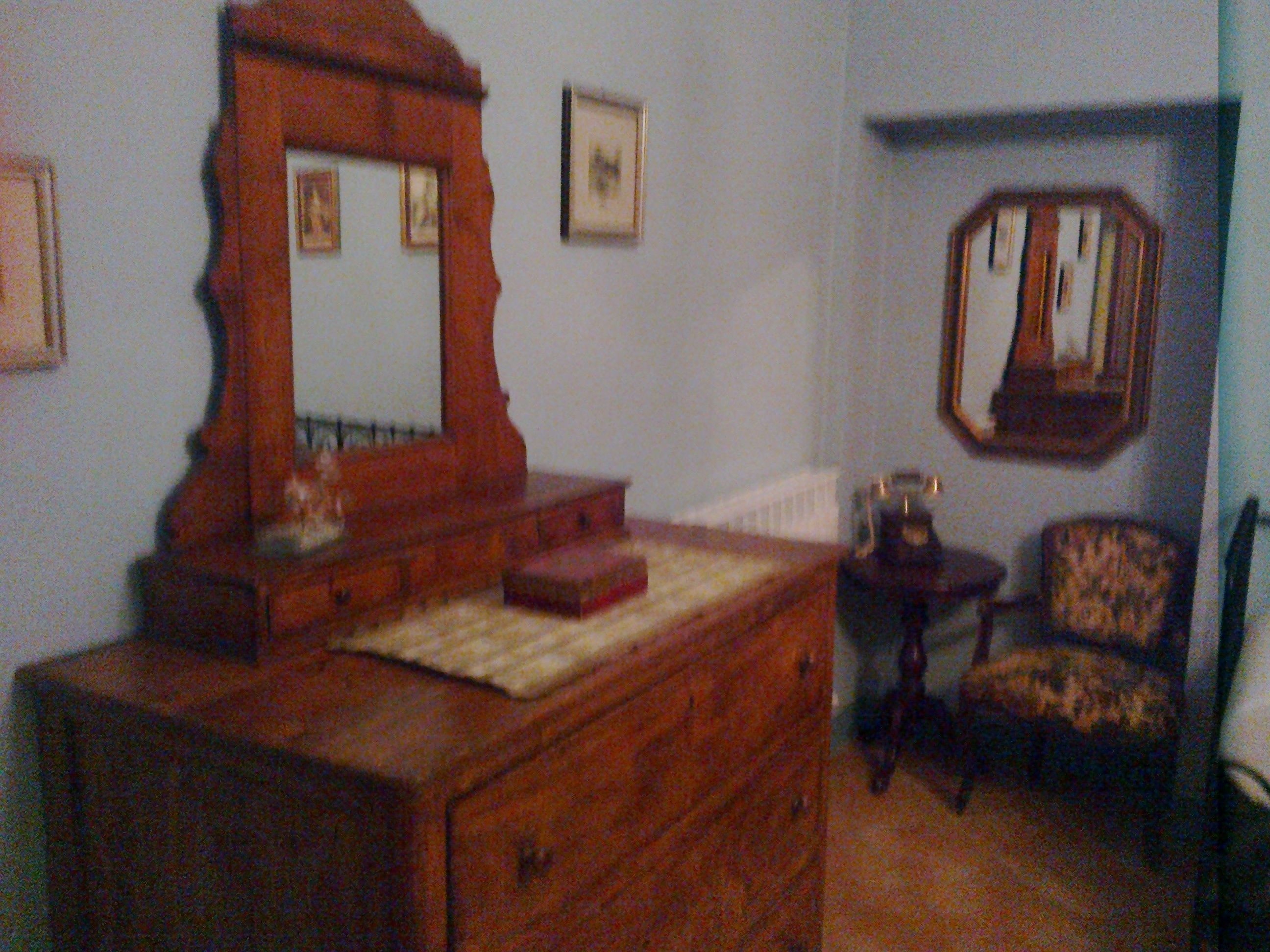 A room with a wooden dresser, a mirror, a chair, and a small table with a lamp.