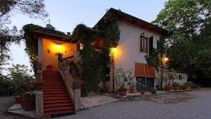 Une maison à deux étages, dotée d’un escalier en pierre menant à l’entrée, entourée de plantes en pot et bordée d’une allée en gravier.