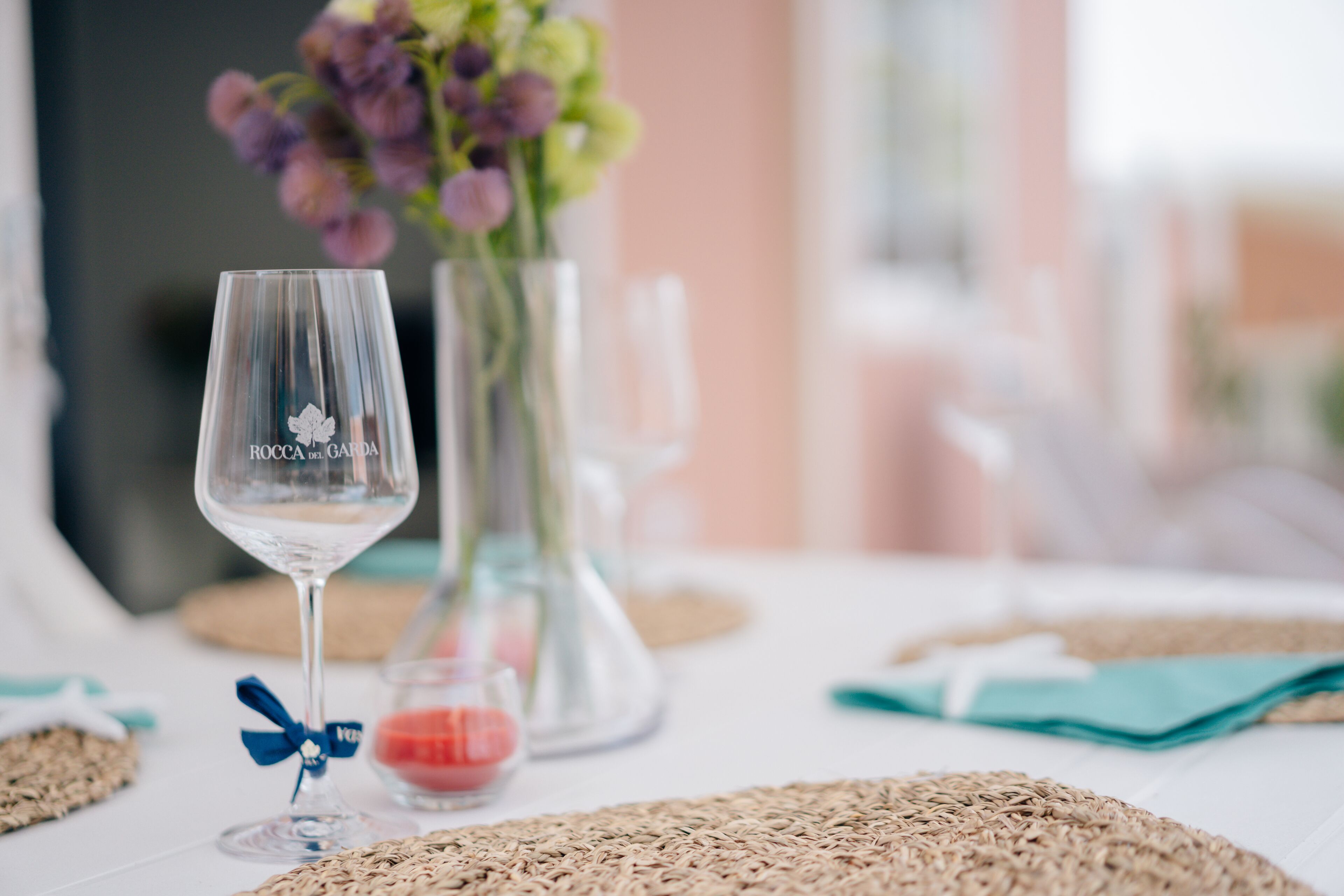 Une table dressée avec un verre à vin transparent, un vase rempli de fleurs et un set de table en fibres tressées.