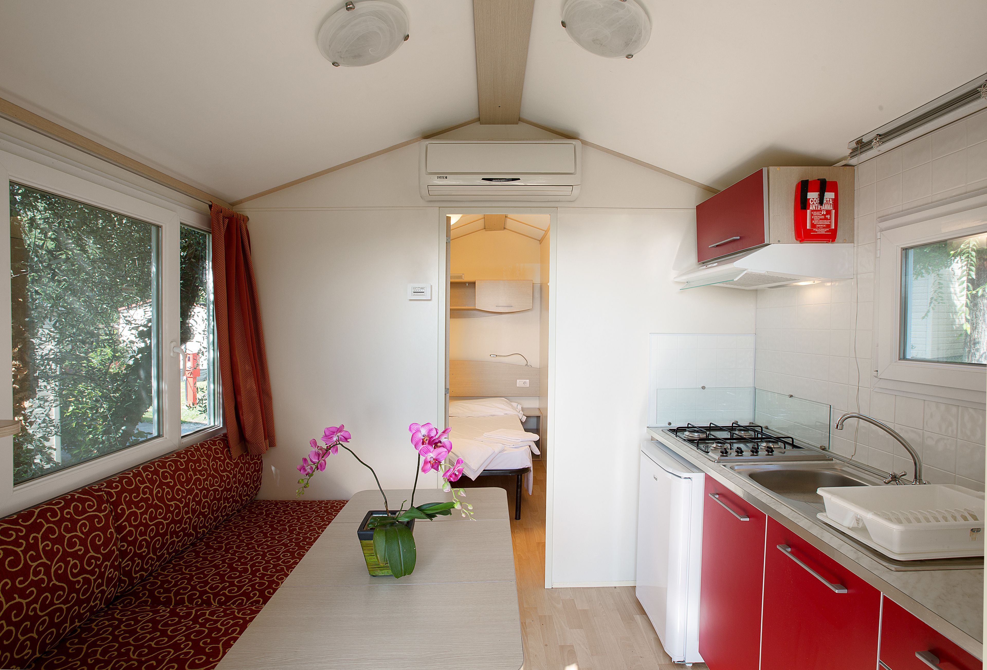 A compact kitchen with a stove, sink, and red cabinets. A window with curtains is on the left, and a bed is visible through an open door in the background.