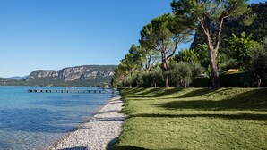 Blick auf einen See mit Kiesstrand, einer sich ins Wasser erstreckenden Mole und einem Hintergrund aus Bergen und Bäumen.