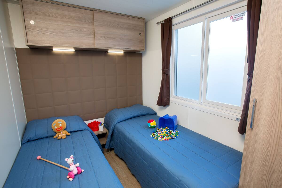 A bedroom with two beds, a window with curtains, a wooden headboard, and a shelf with toys.