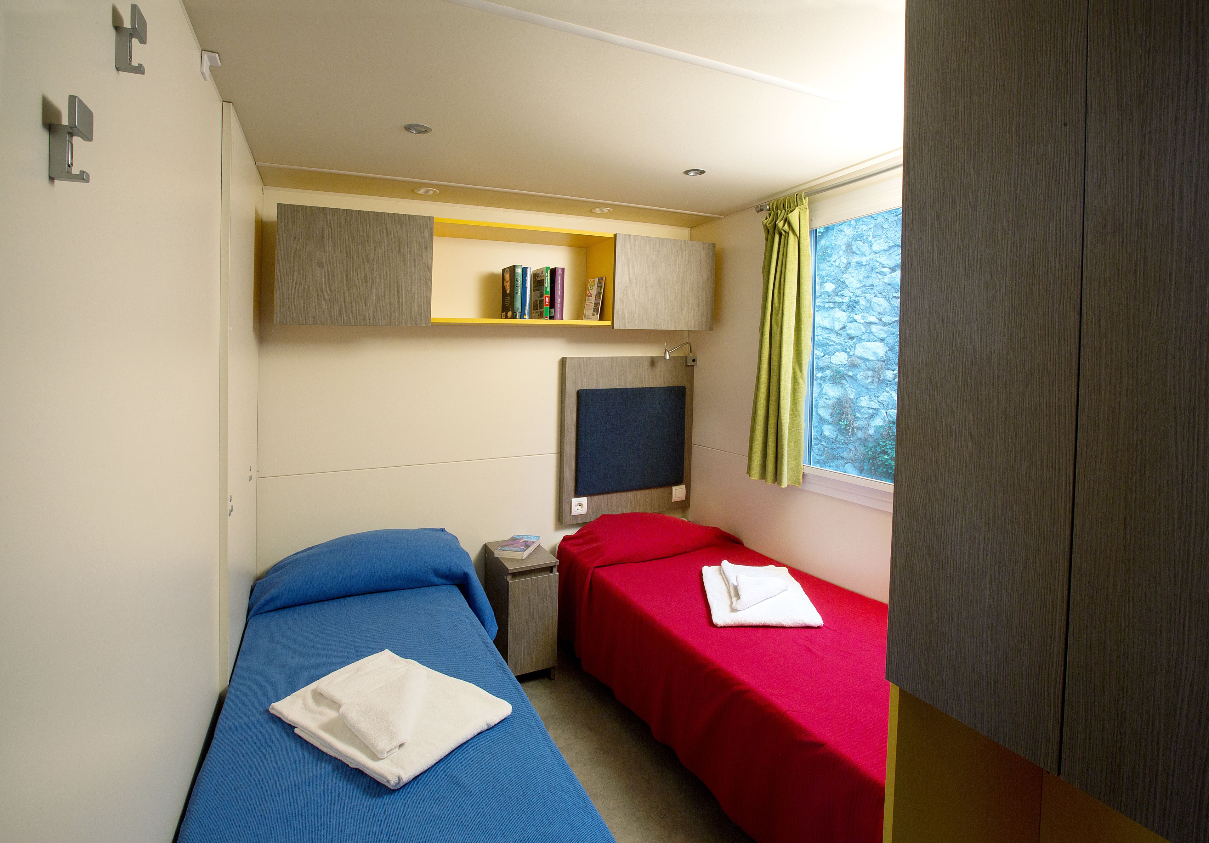 A small hotel room with two beds, a TV, a shelf with books, and a window with curtains.